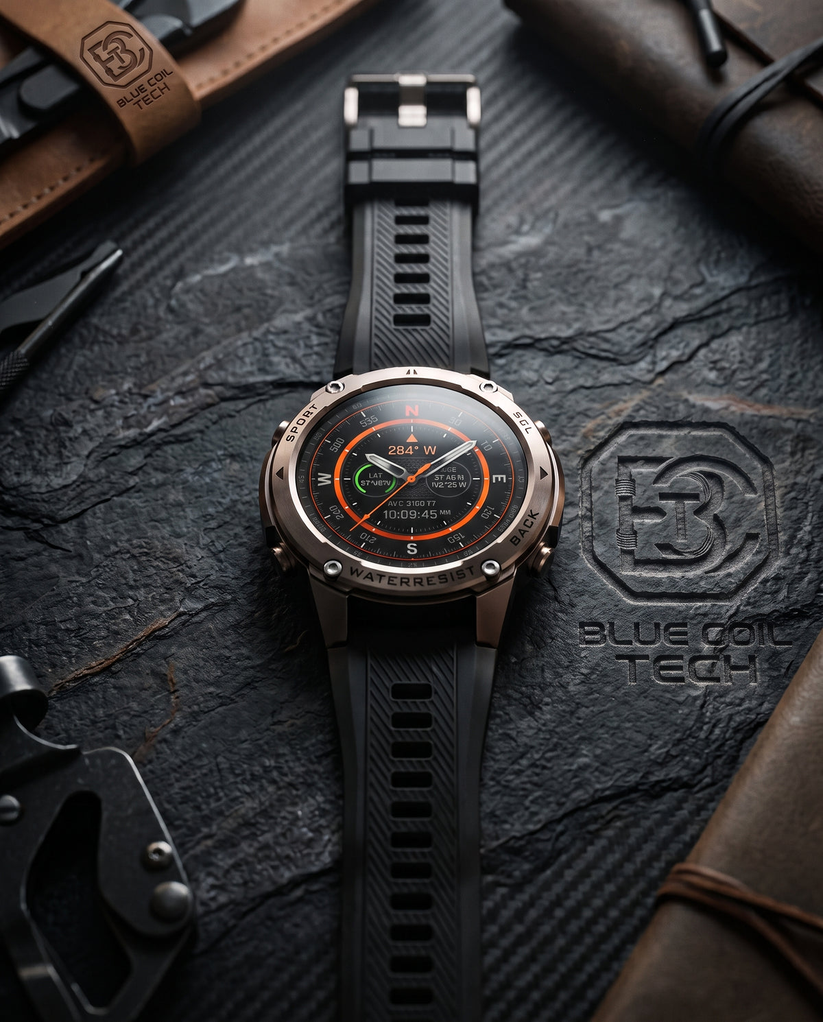 A rugged smartwatch with a black textured strap and a bronze-toned case, displayed on a dark textured surface with leather accessories and branded Blue Coil Tech branding.